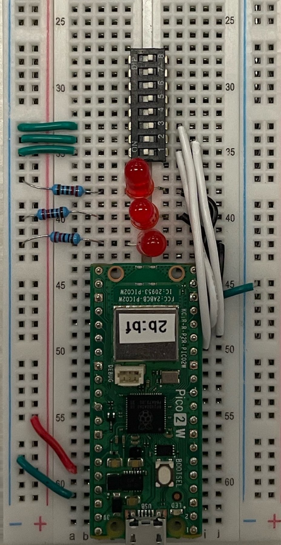 A Pico 2 W breadboard circuit with three red LEDs wired to GP13, GP14, and GP15, and an 8-position DIP switch added above them straddling the center gap. Three of its switches have a green wire running to the main ground rail and a white wire running to the Pico's GP10, GP11, and GP12 pins.